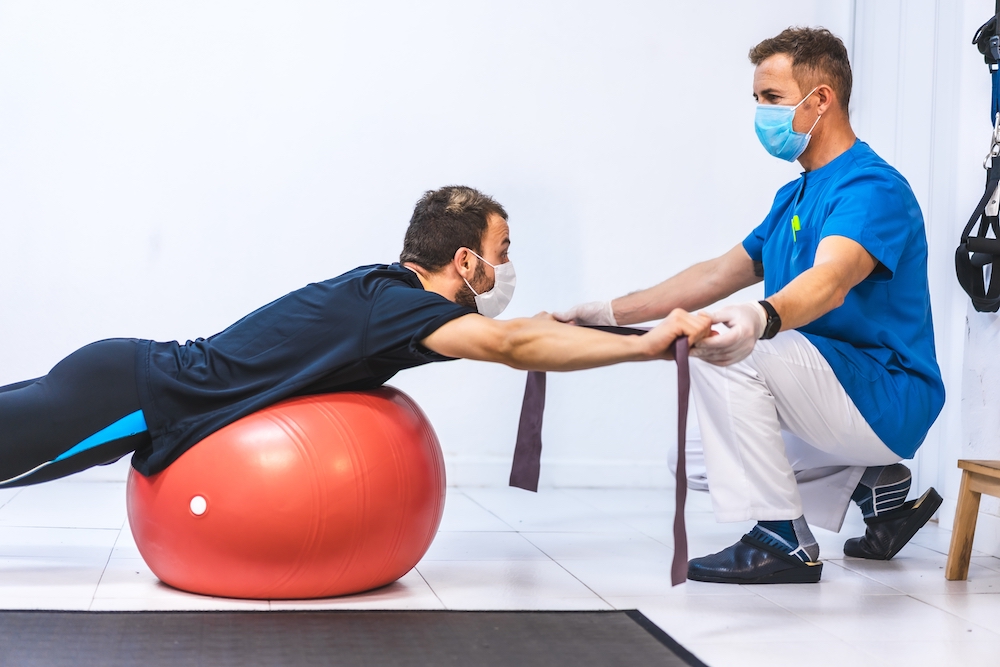A physiotherapist with medical mask helping to exercise to a male with a black shirt